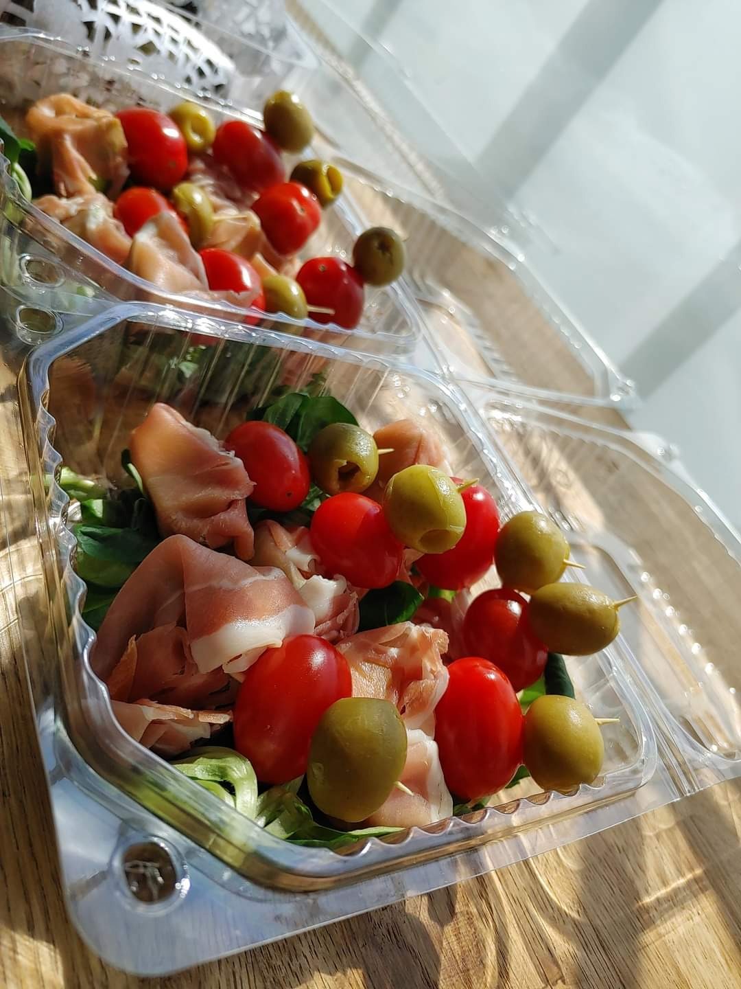 Dwa przezroczyste plastikowe pojemniki z sałatką: zielone liście, pomidorki cherry, plasterki szynki parmeńskiej i zielone oliwki na wykałaczkach, ustawione na drewnianym stole w jasnym świetle.