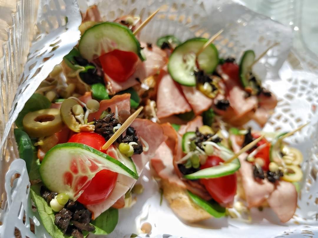 Dekoracyjne, białe opakowanie z kanapkami na jeden kęs: pieczywo, szynka, pomidory, ogórki, oliwki, kiełki, spięte wykałaczkami.