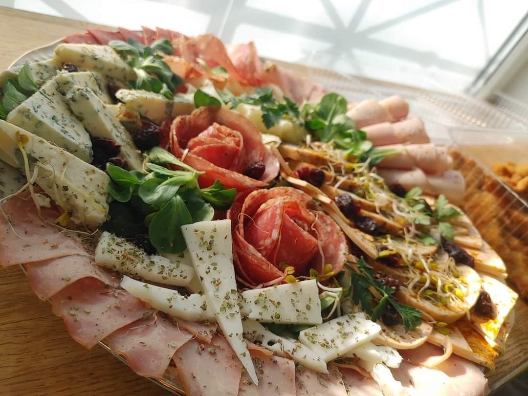 Dekoracyjny talerz z przekąskami: wędliny ułożone w rozety, sery pleśniowe i białe posypane ziołami, bagietki z pastą i kiełkami, żurawina, zielenina, oświetlone naturalnym światłem z okna.