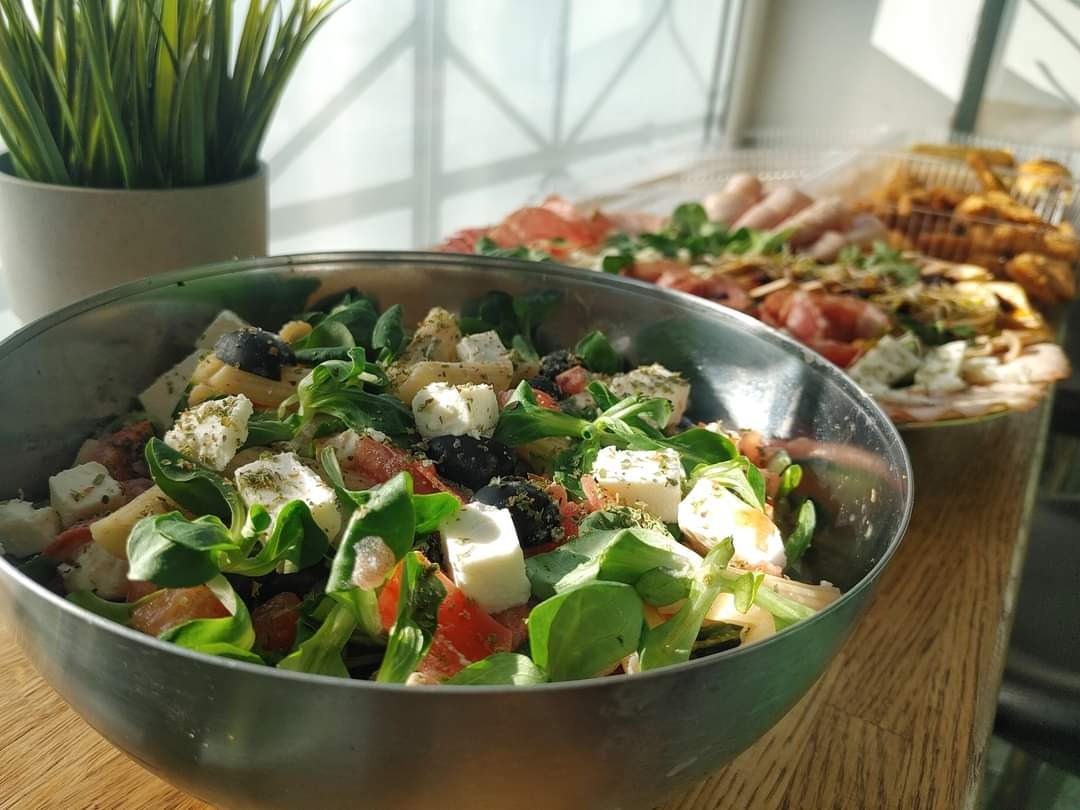 Metalowa misa z sałatką z pomidorów, sera feta, oliwek i roszponki, posypana ziołami, na drewnianym blacie, z widocznymi w tle tacami z przekąskami i rośliną doniczkową.