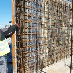 Zbrojenie pionowej konstrukcji żelbetowej w szalunku, widoczne elementy stalowe i betonowe. Pracownik w rękawicach i kamizelce odblaskowej przy szalunku.