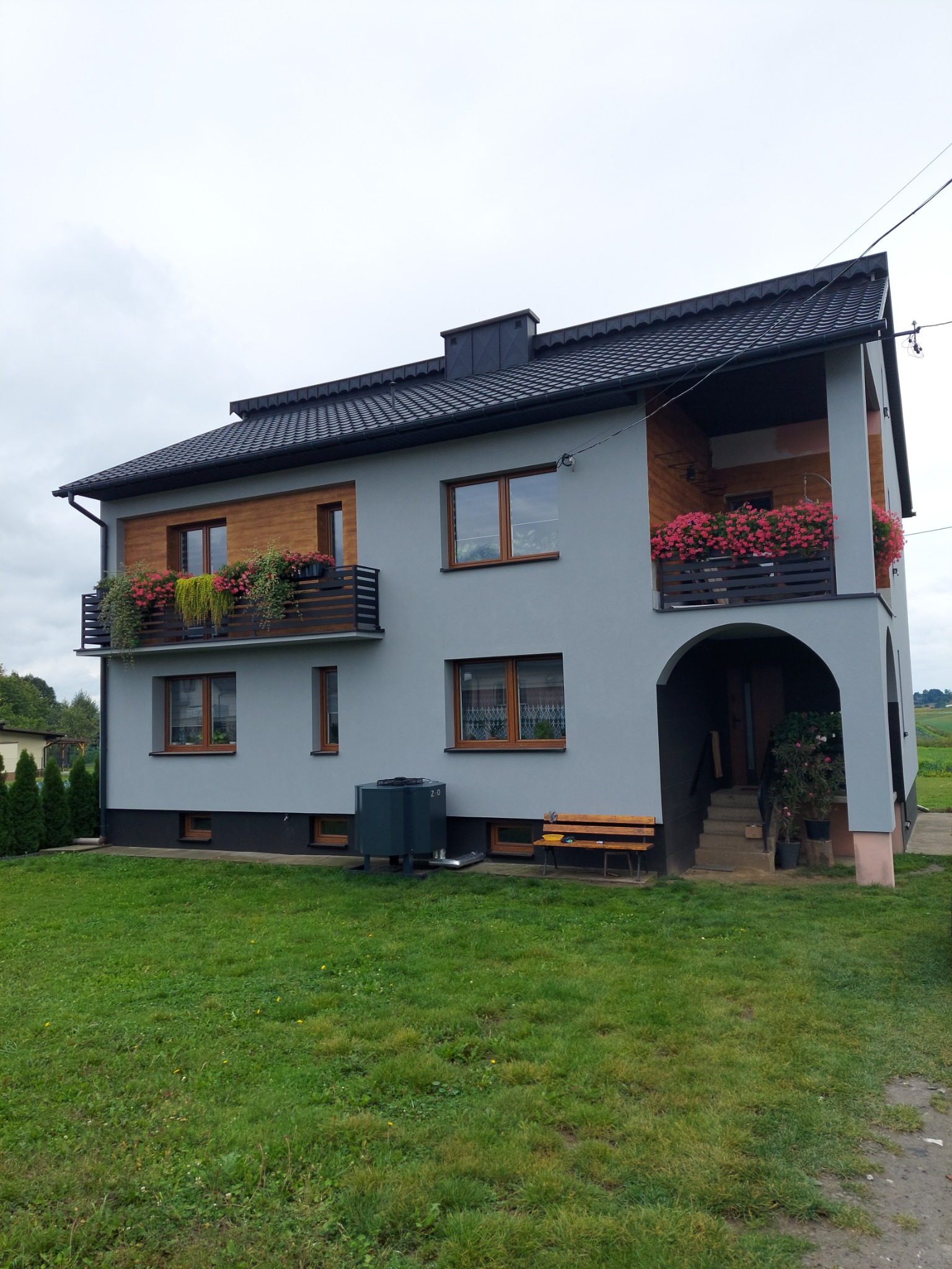 Dwupiętrowy dom z szarą elewacją, drewnianymi akcentami i balkonami ozdobionymi kwiatami, widoczny z perspektywy trawnika, z zainstalowanym urządzeniem na zewnątrz.