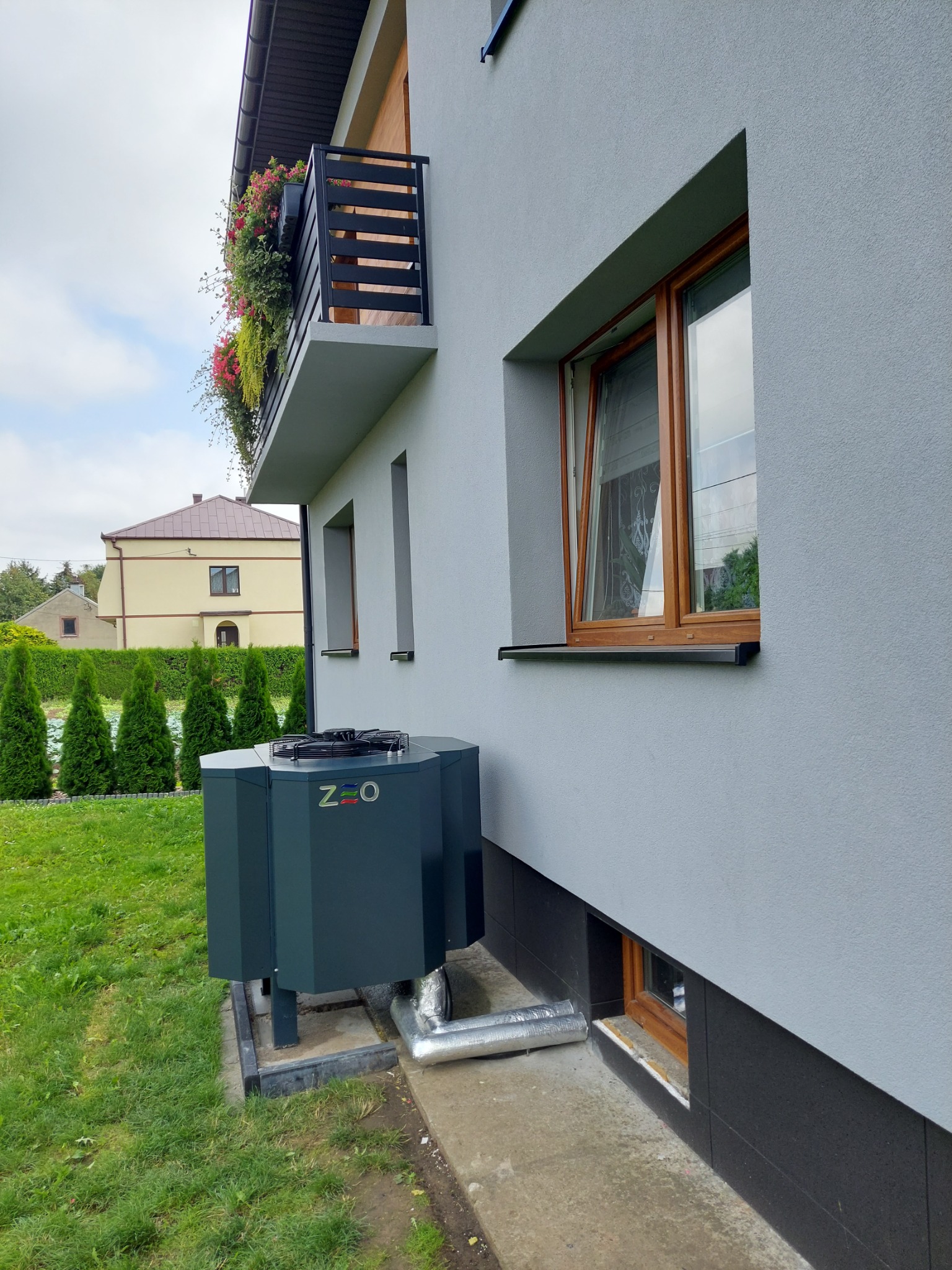 Szara pompa ciepła przy ścianie domu z oknami i balkonem z kwiatami, widoczne rury przyłączeniowe.