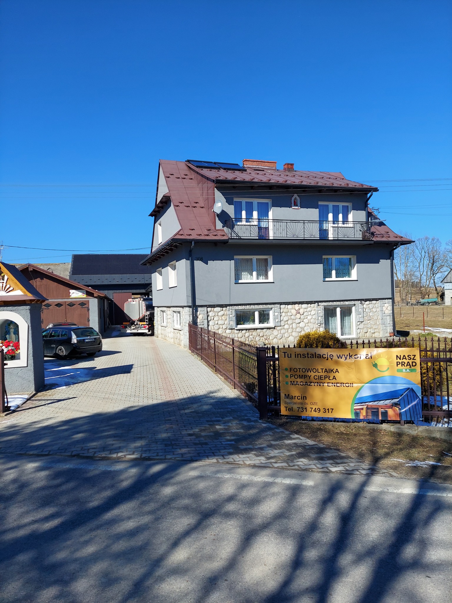 Dom z panelami fotowoltaicznymi na dachu, widoczny znak reklamowy firmy instalującej fotowoltaikę, pompy ciepła i magazyny energii.