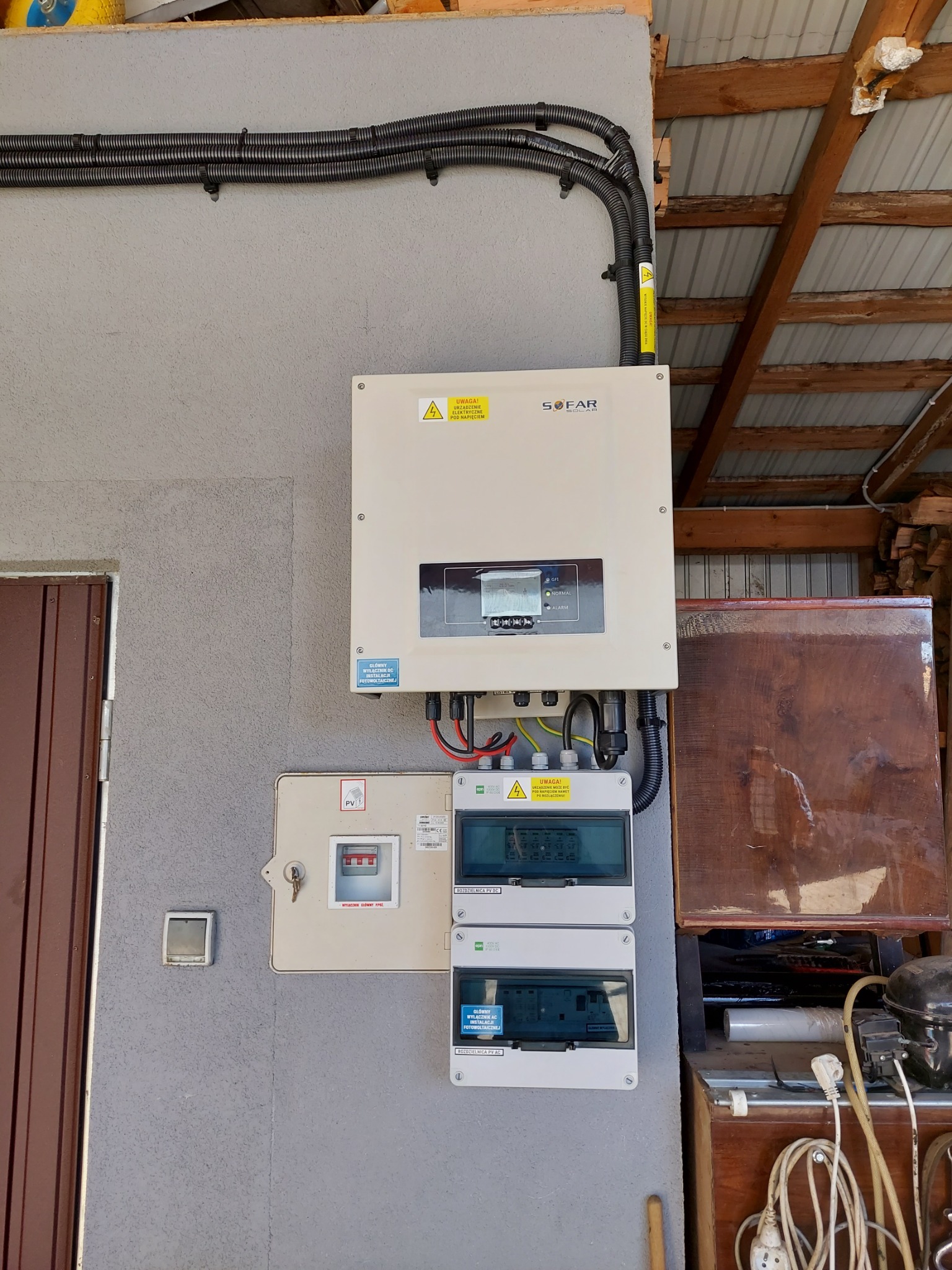 Instalacja z inwerterem SOFAR i rozdzielnicami elektrycznymi na ścianie budynku w Ładnej, z widocznymi przewodami w peszlach i tabliczkami ostrzegawczymi.