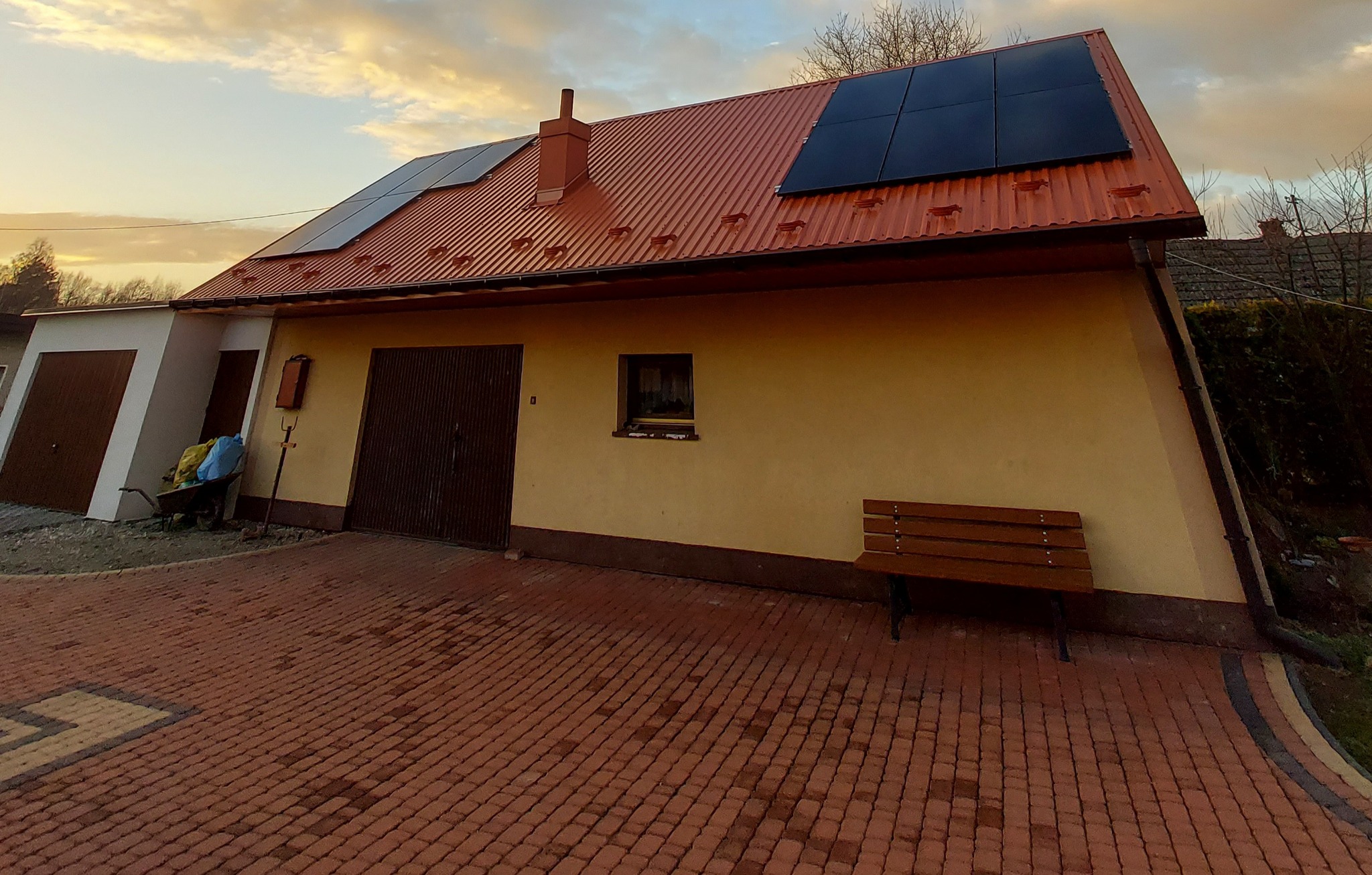 Dom jednorodzinny z czerwonym dachem, na którym zamontowano panele słoneczne; widoczny podjazd z kostki brukowej i drewniana ławka przy ścianie budynku.