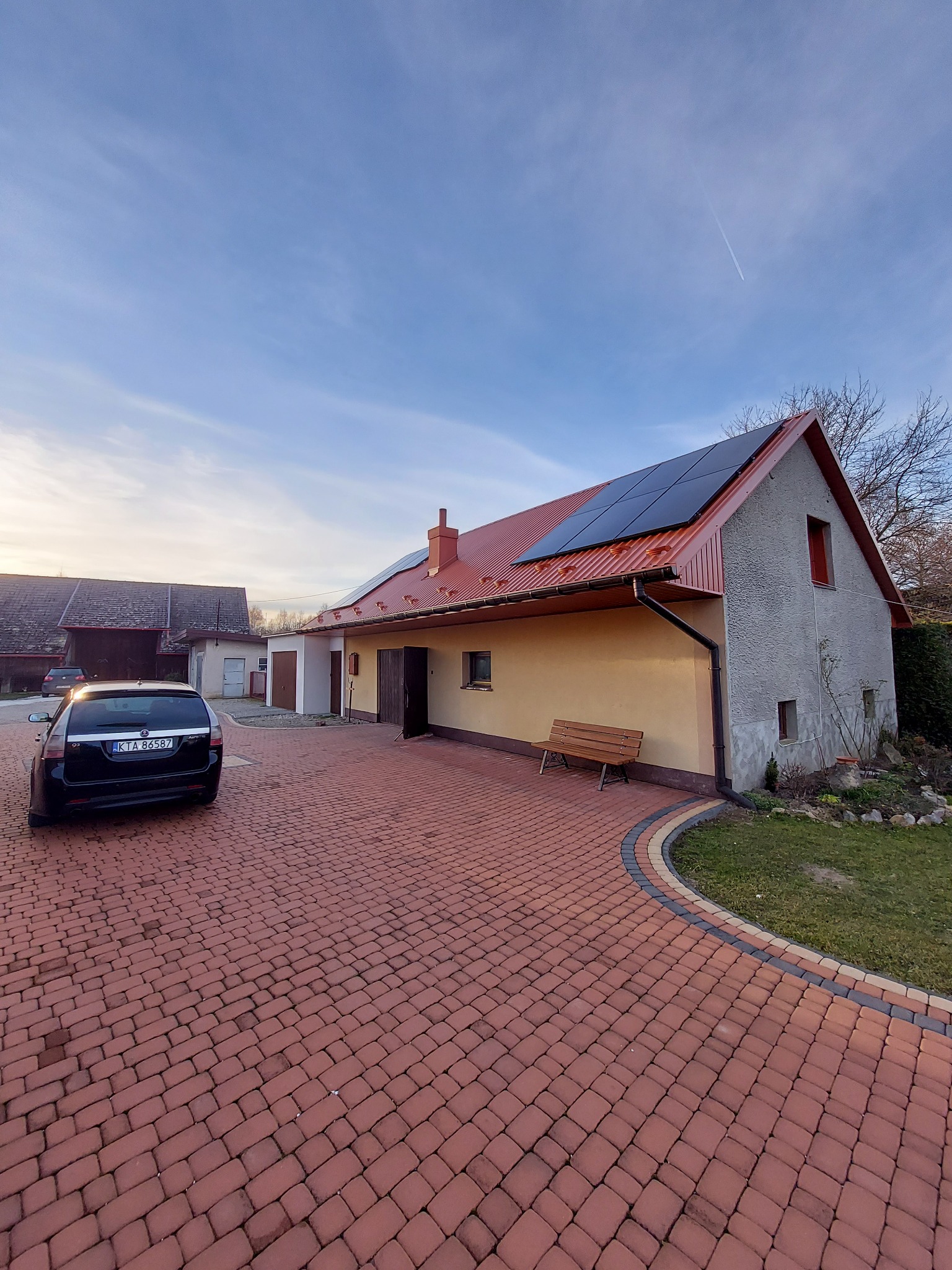Dom jednorodzinny z panelami słonecznymi na czerwonym dachu, widok z brukowanego podjazdu, na pierwszym planie zaparkowany czarny samochód.