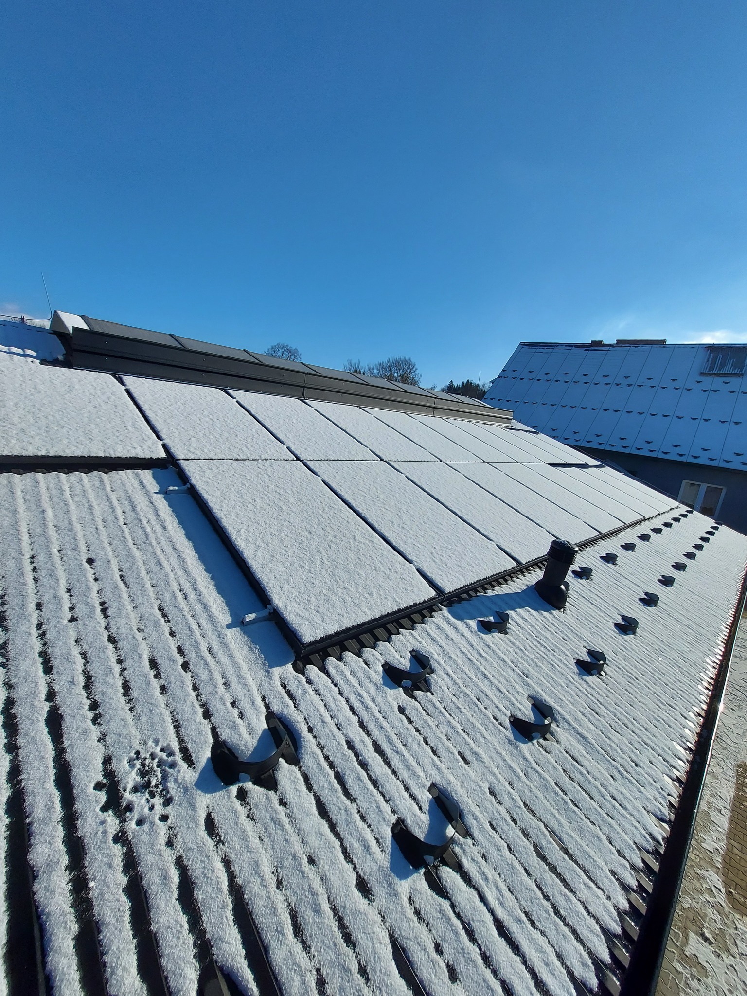 Panel słoneczny pokryty śniegiem na dachu budynku w słoneczny dzień, widoczne elementy montażowe i fragment dachu sąsiedniego budynku.