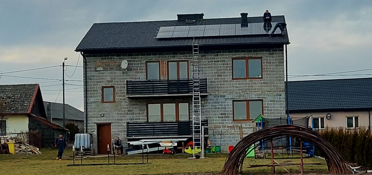 Montaż paneli słonecznych na dachu dwupiętrowego domu z szarej cegły, drabina oparta o dach, dwóch instalatorów na dachu, pochmurne niebo.