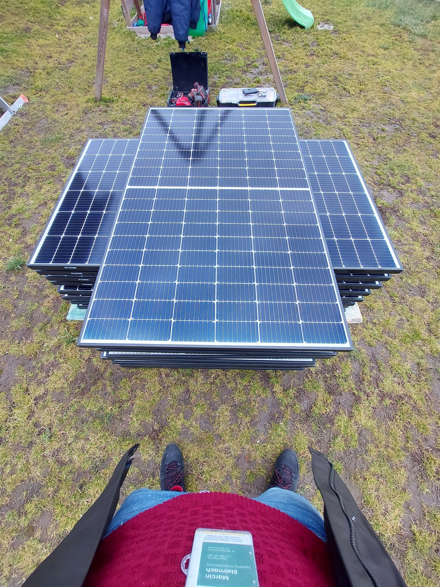 Stos paneli fotowoltaicznych ułożonych na trawie przed montażem, widok z góry z perspektywy osoby trzymającej telefon z widoczną wizytówką firmy fotowoltaicznej.