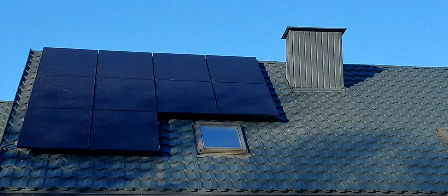 Czarny panel fotowoltaiczny zamontowany na szarym dachu z dachówki ceramicznej, obok komina i okna dachowego, na tle błękitnego nieba.