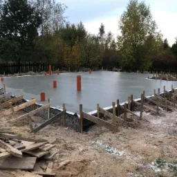 Wylana płyta fundamentowa z wystającymi pionowymi rurami kanalizacyjnymi, widoczne szalunki i drewniane podpory, w tle drzewa i ogrodzenie.