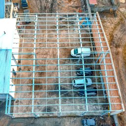 Stalovers - Widok z góry na konstrukcję stalową w trakcie budowy, z zaparkowanymi samochodami wewnątrz szkieletu, niebieskie plandeki zabezpieczające na niektórych elementach.