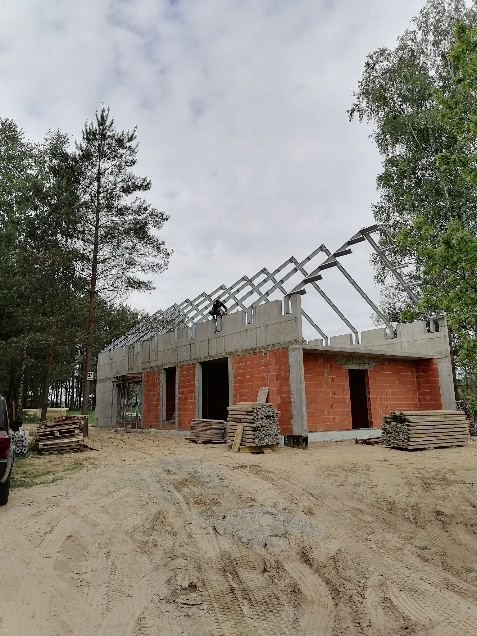 Budowa domu w stanie surowym otwartym: ceglano-betonowe ściany, metalowa konstrukcja dachu, pracownik na dachu, stosy drewna obok budynku, piaszczyste podłoże.