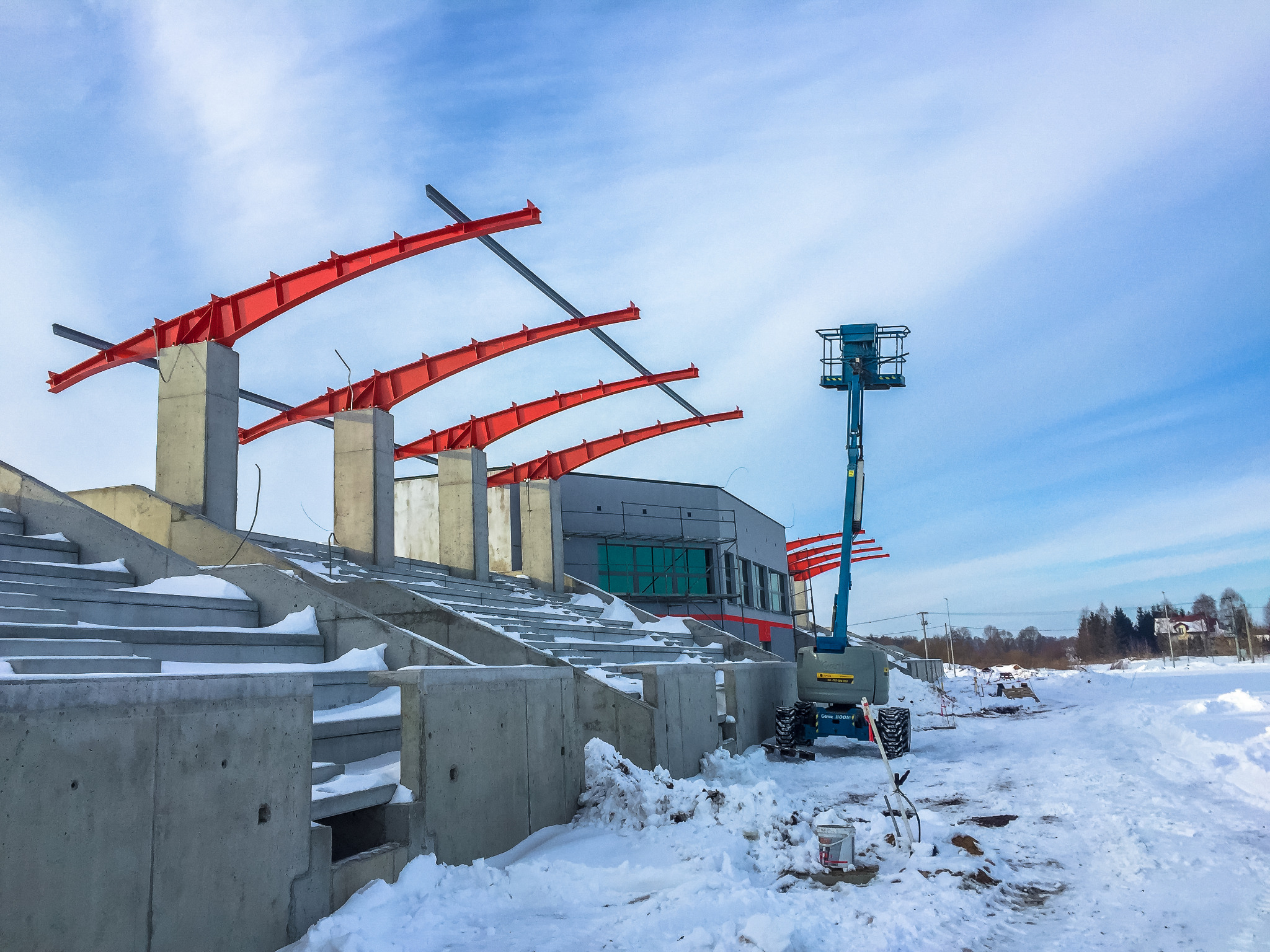 Budowa betonowych trybun sportowych w zimowej scenerii, widoczny dźwig platformowy i czerwone elementy konstrukcyjne dachu.