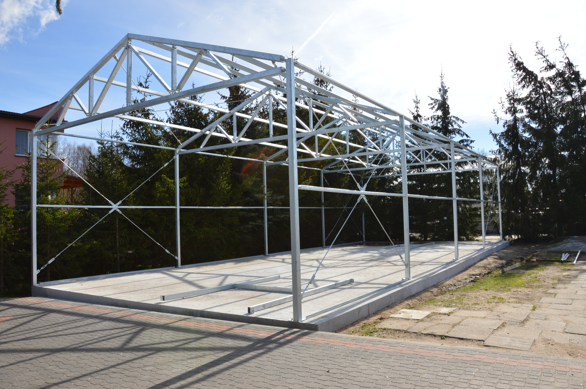 Metalowa konstrukcja szkieletowa domu na betonowej płycie fundamentowej, widok z zewnątrz, drzewa w tle.