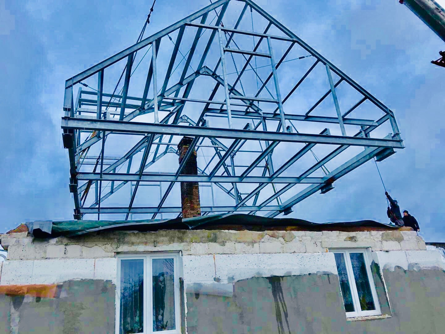 Montaż stalowej konstrukcji dachu na istniejącym budynku z kominem, widoczne okna i pracownicy zabezpieczający konstrukcję