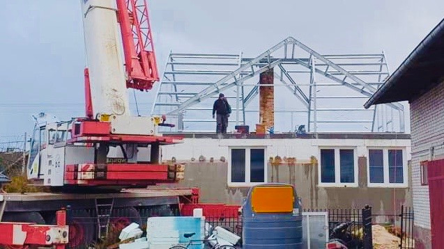 Budowa domu z widoczną konstrukcją szkieletową dachu, dźwigiem i pracownikiem na górze, w trakcie montażu.