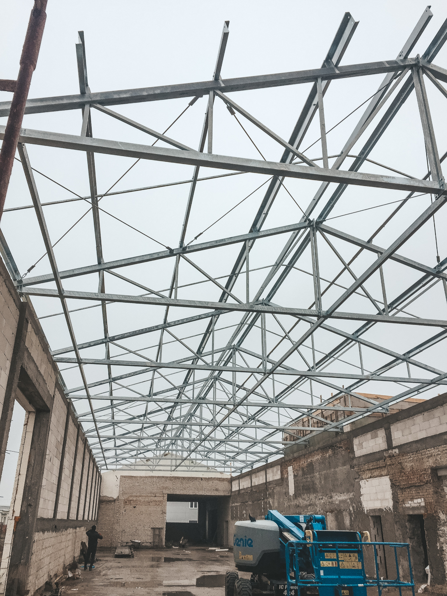 Konstrukcja stalowa dachu w trakcie budowy, widoczny podnośnik koszowy Genie i fragmenty ścian z cegły oraz bloczków betonowych, pochmurne niebo w tle.
