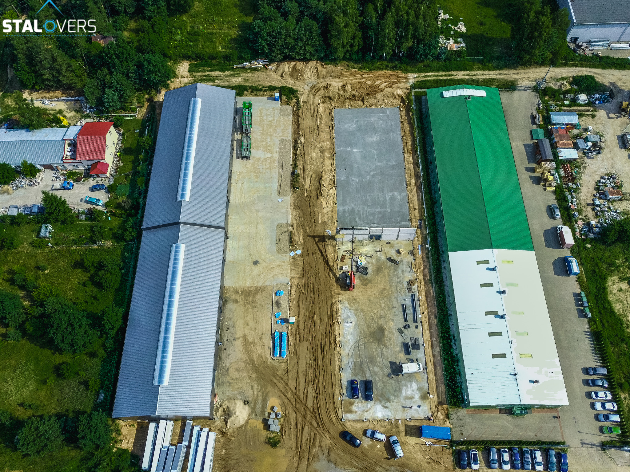 Widok z lotu ptaka na plac budowy z trzema halami: szarą z długimi świetlikami, zieloną i betonową płytą fundamentową w trakcie przygotowania, widoczne materiały budowlane, maszyny i zaparkowane...