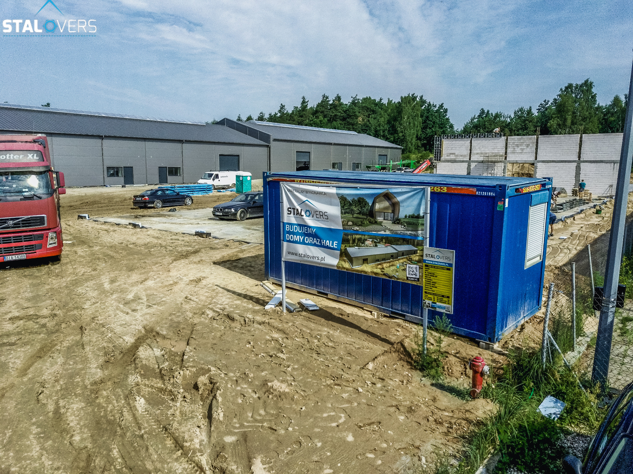 Plac budowy z kontenerem biurowym, na którym umieszczono baner reklamowy firmy budowlanej, w tle budynek hali i wznoszone mury z betonu komórkowego.