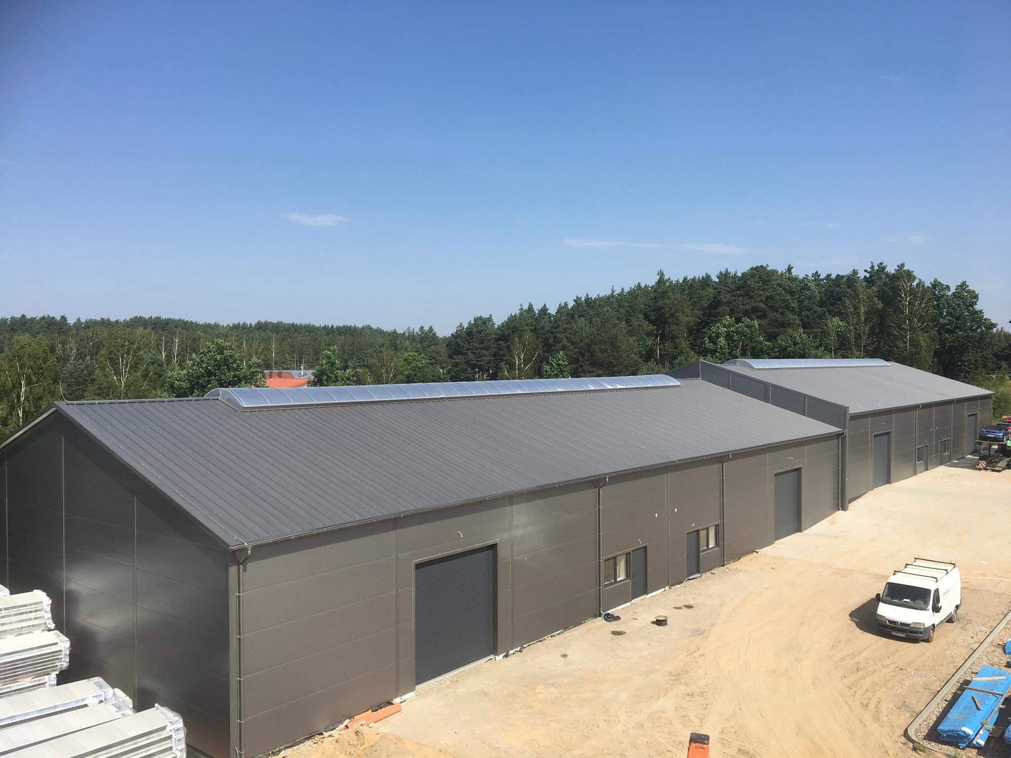 Dwa hale magazynowe z szarej blachy trapezowej, widoczne panele solarne na dachu, samochód dostawczy na placu budowy.