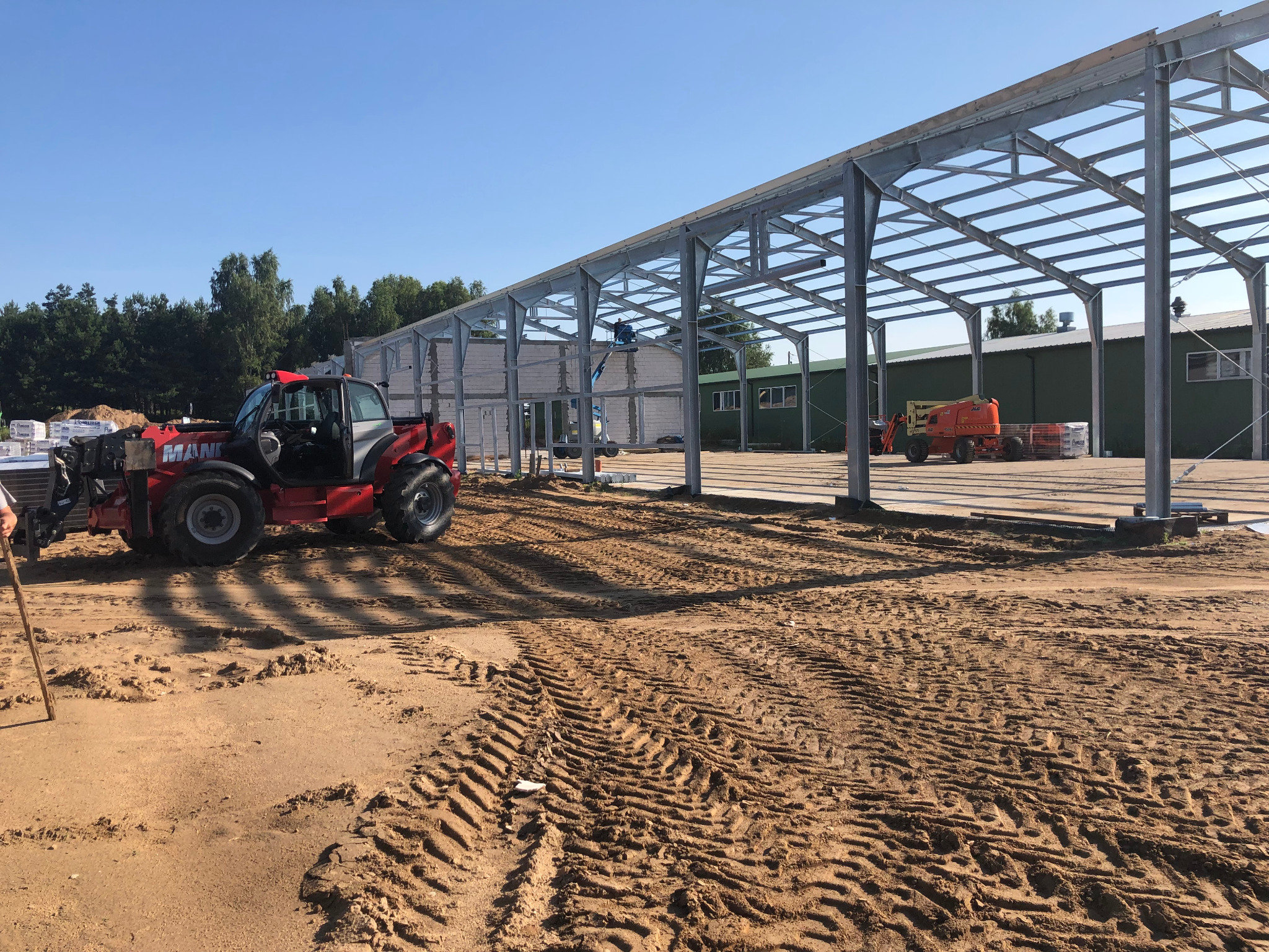 Montaż konstrukcji stalowej hali z użyciem podnośników teleskopowych i ładowarki teleskopowej Manitou, widoczne ślady opon na piaszczystym podłożu.