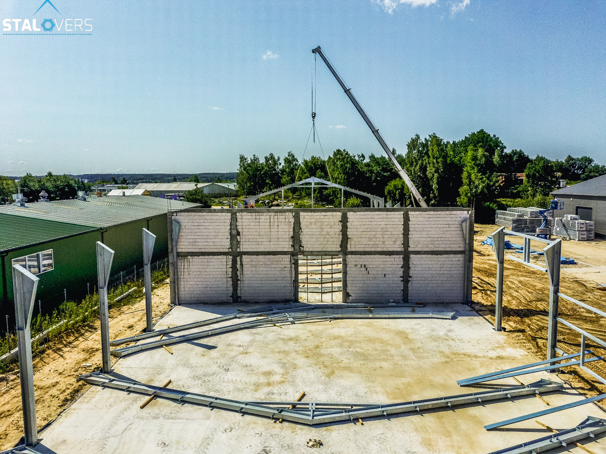 Budowa hali: wznoszone ściany z betonu komórkowego i stalowa konstrukcja dachu podnoszona przez dźwig na placu budowy.