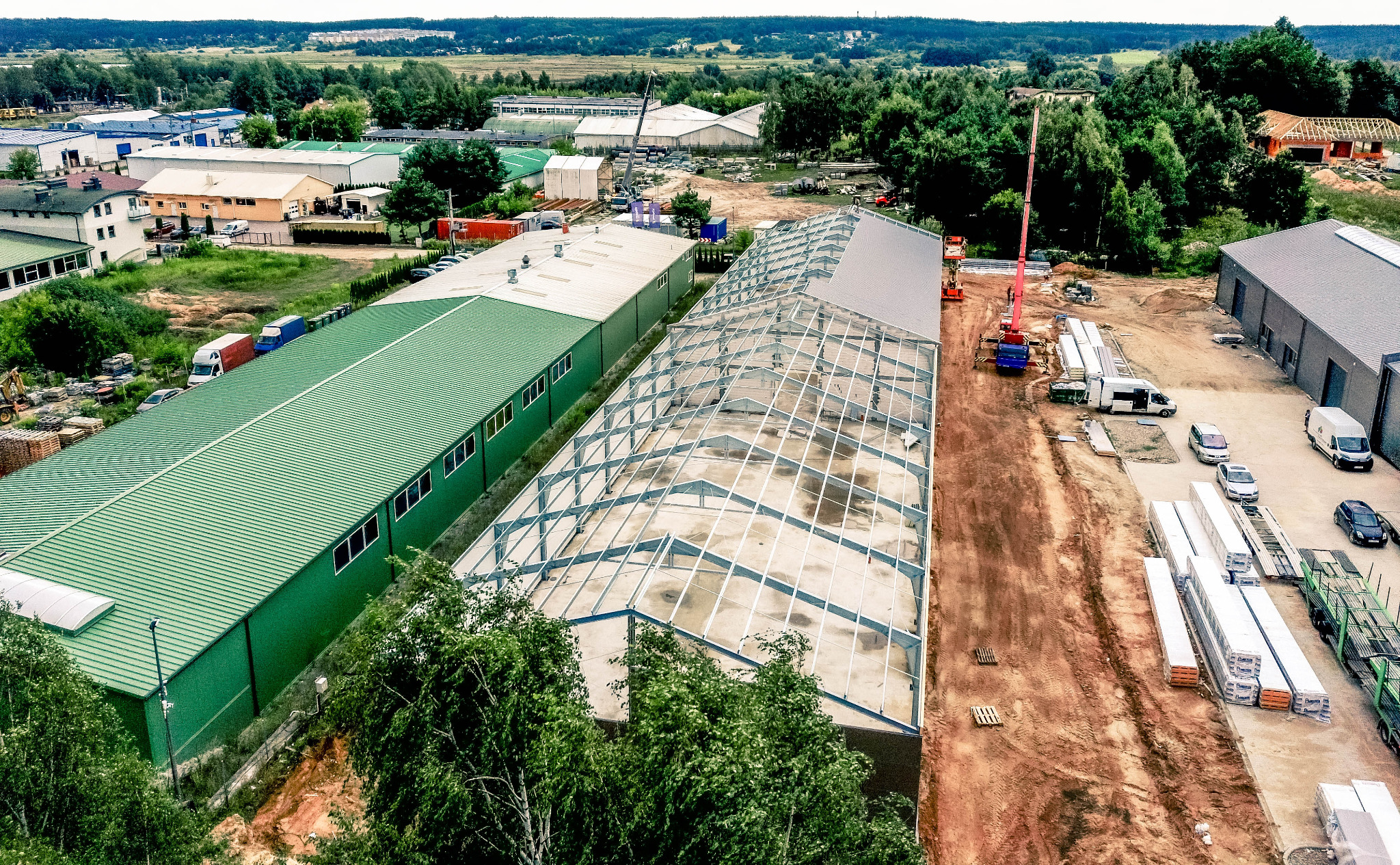 Widok z góry na budowę hali z metalową konstrukcją szkieletową, obok gotowy budynek z zielonym dachem, w tle las i zabudowania, na placu budowy dźwig i materiały budowlane.