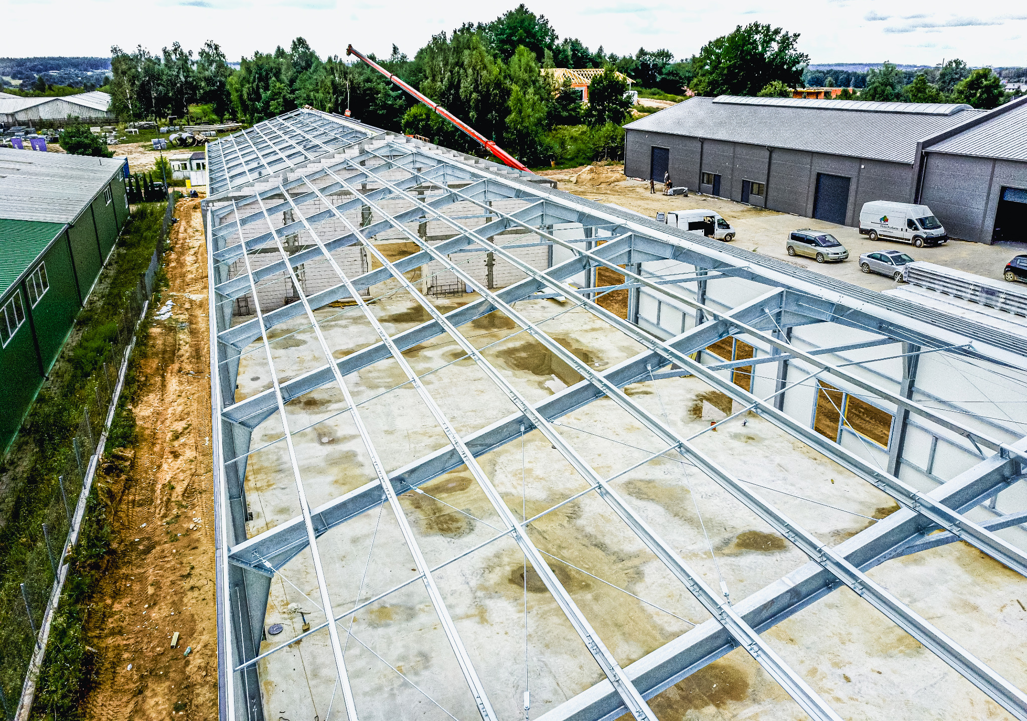 Widok z lotu ptaka na budowę hali z metalową konstrukcją szkieletową, na betonowej płycie fundamentowej, z dźwigiem w tle i budynkami magazynowymi w otoczeniu.