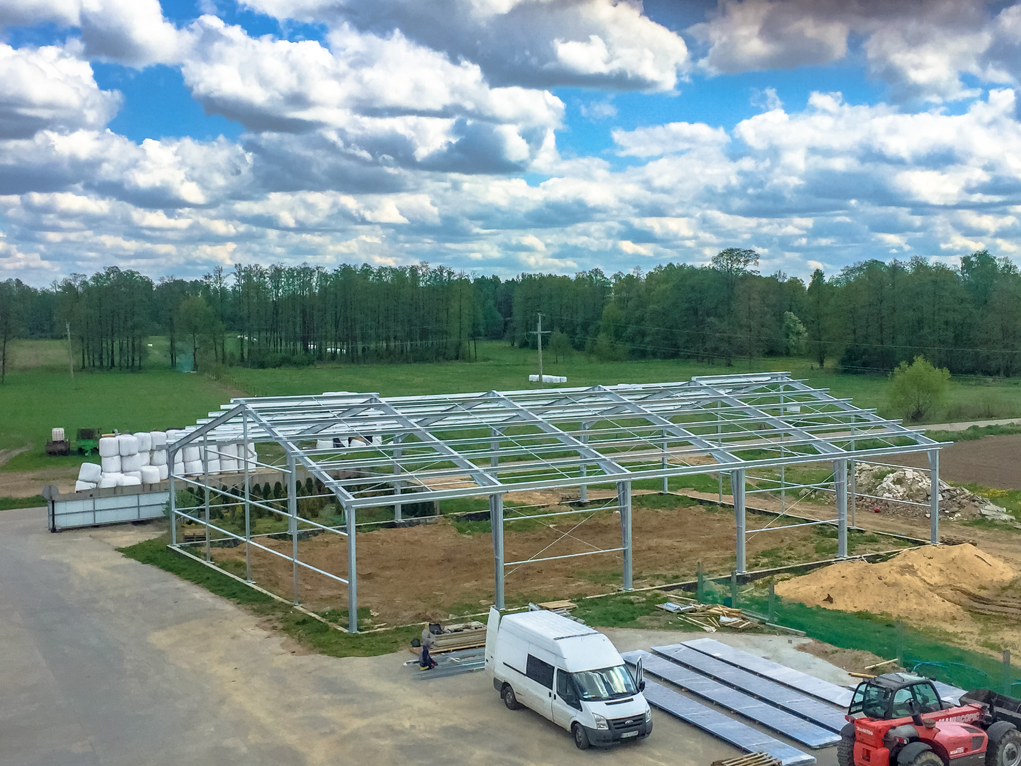 Konstrukcja szkieletowa w trakcie budowy na tle pola i lasu, z widocznymi materiałami budowlanymi, samochodem dostawczym i czerwoną ładowarką teleskopową.
