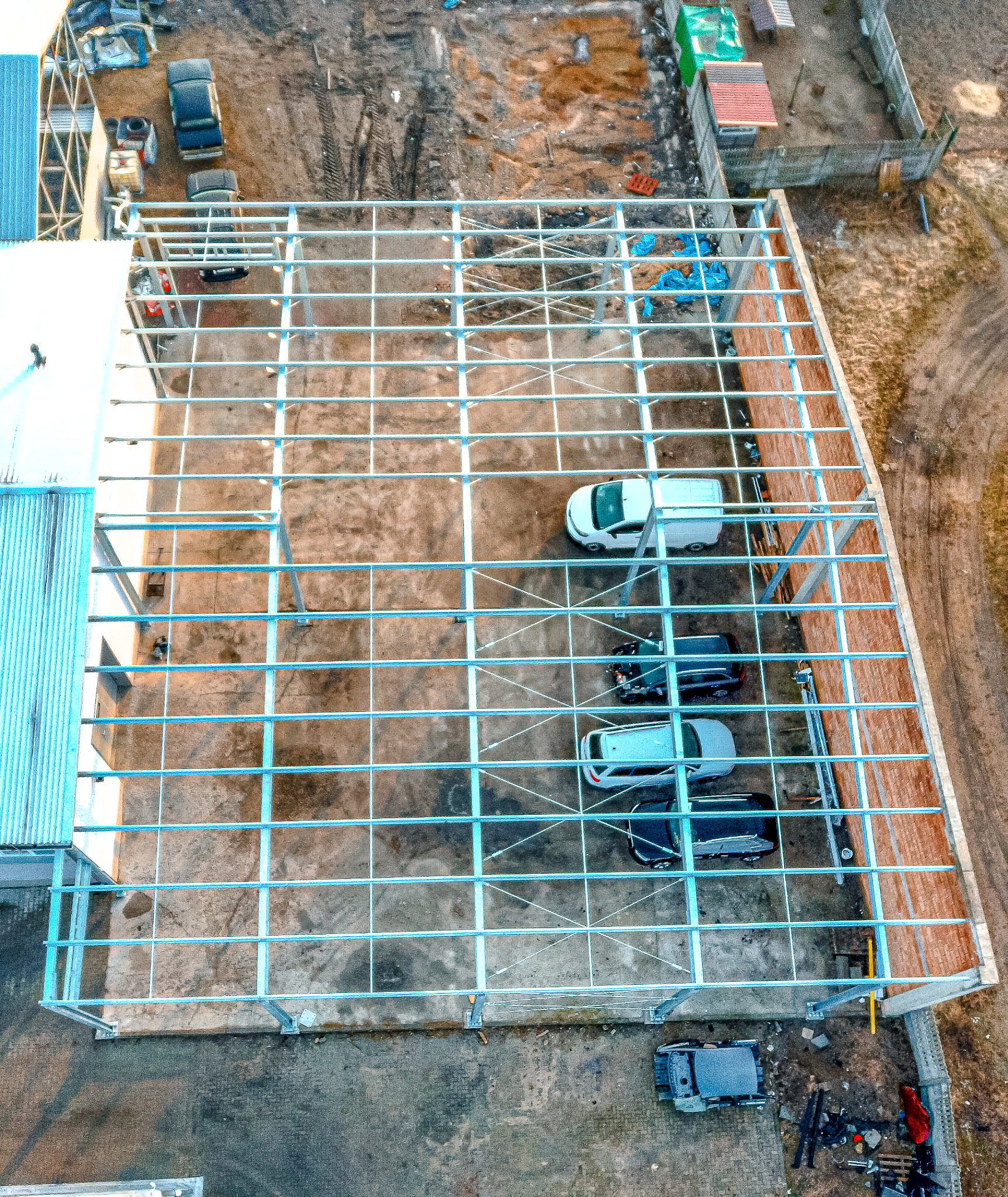 Widok z góry na konstrukcję stalową w trakcie budowy, z zaparkowanymi samochodami wewnątrz szkieletu, niebieskie plandeki zabezpieczające na niektórych elementach.
