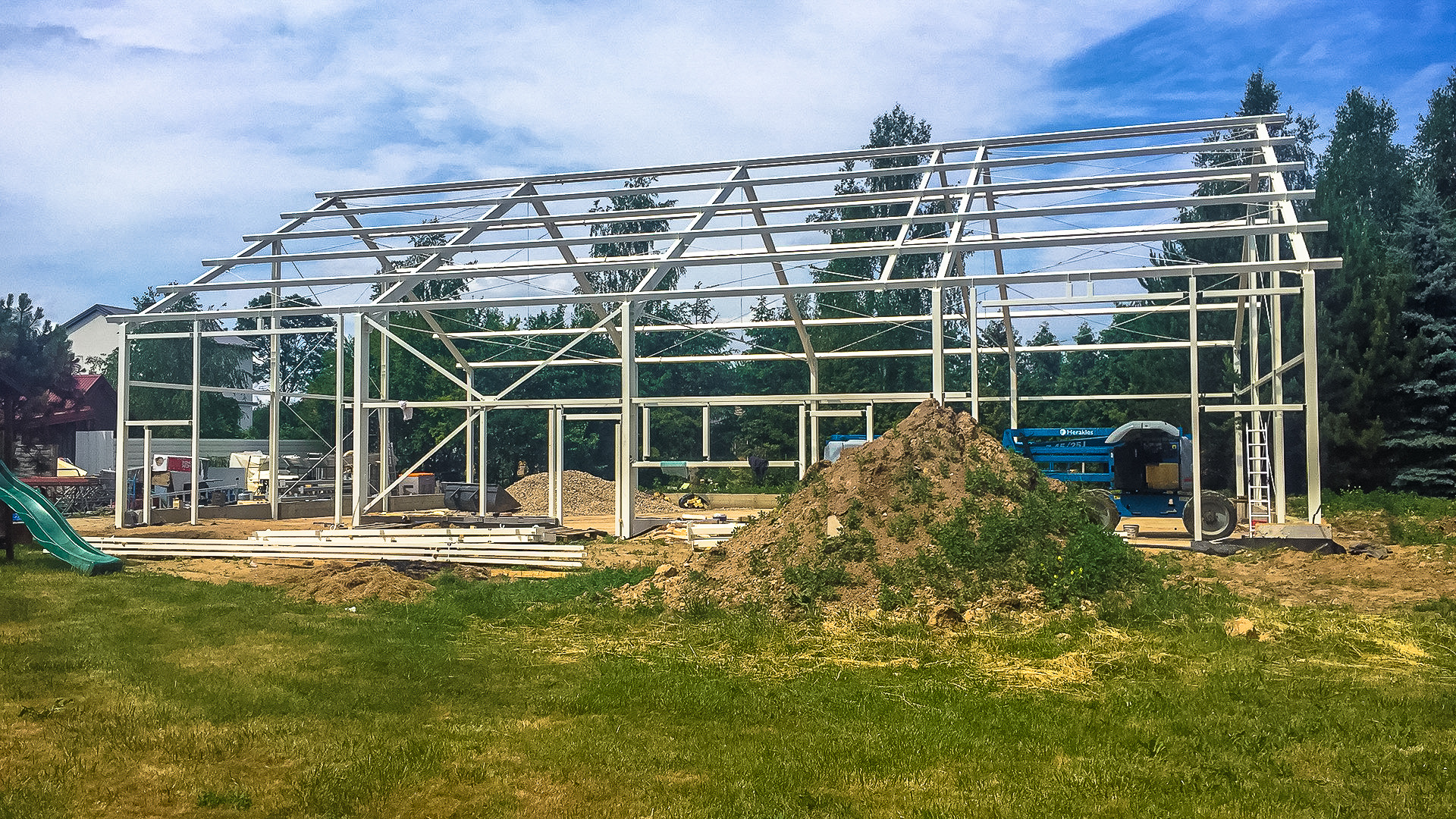 Konstrukcja szkieletowa domu w trakcie montażu, widoczny podnośnik koszowy Herakles, pryzma ziemi i zjeżdżalnia dla dzieci w tle.