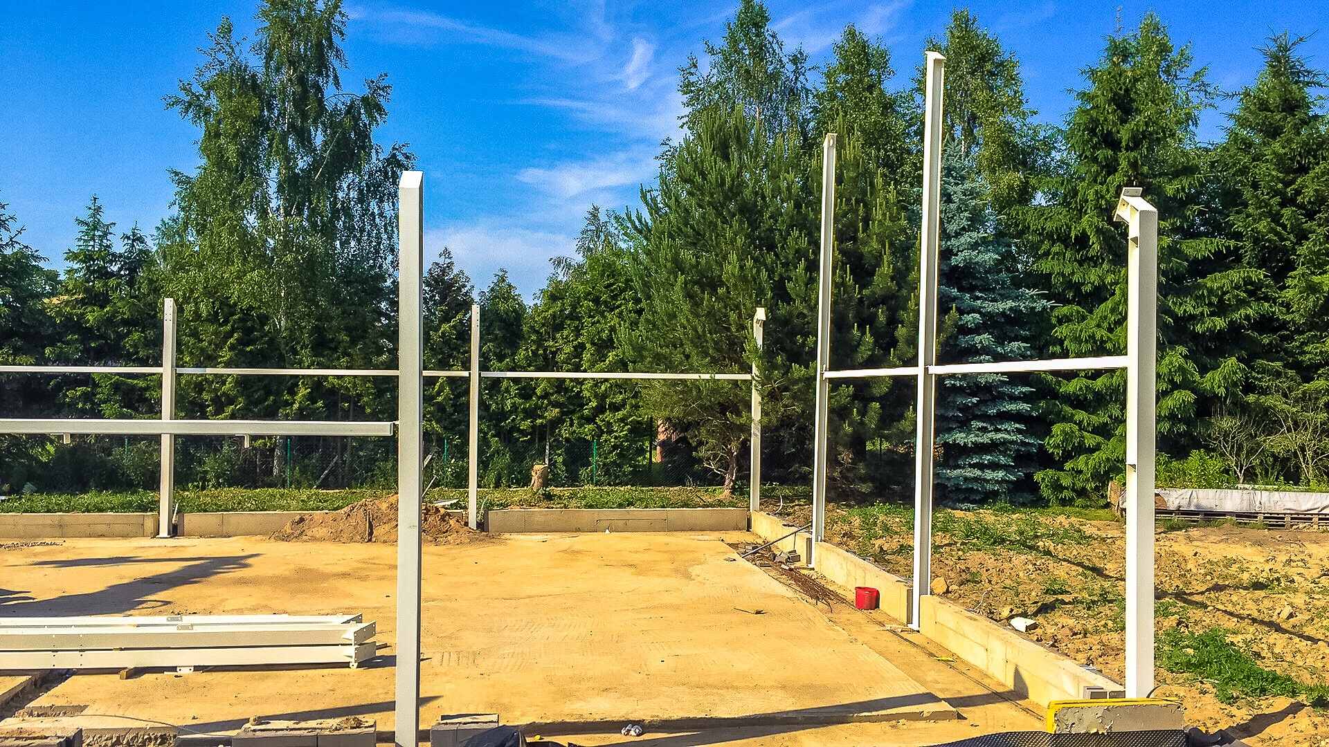 Wczesny etap budowy domu: metalowy szkielet konstrukcyjny na betonowym fundamencie, w tle zielone drzewa i błękitne niebo.