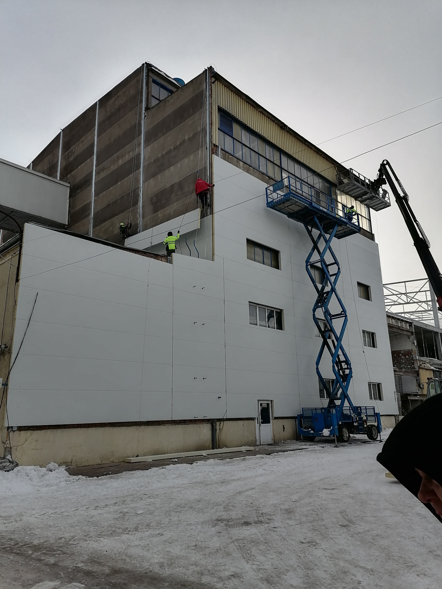 Montaż paneli elewacyjnych na budynku przemysłowym z wykorzystaniem podnośnika koszowego i technik alpinistycznych, widoczne fragmenty starej elewacji i śnieg na ziemi.