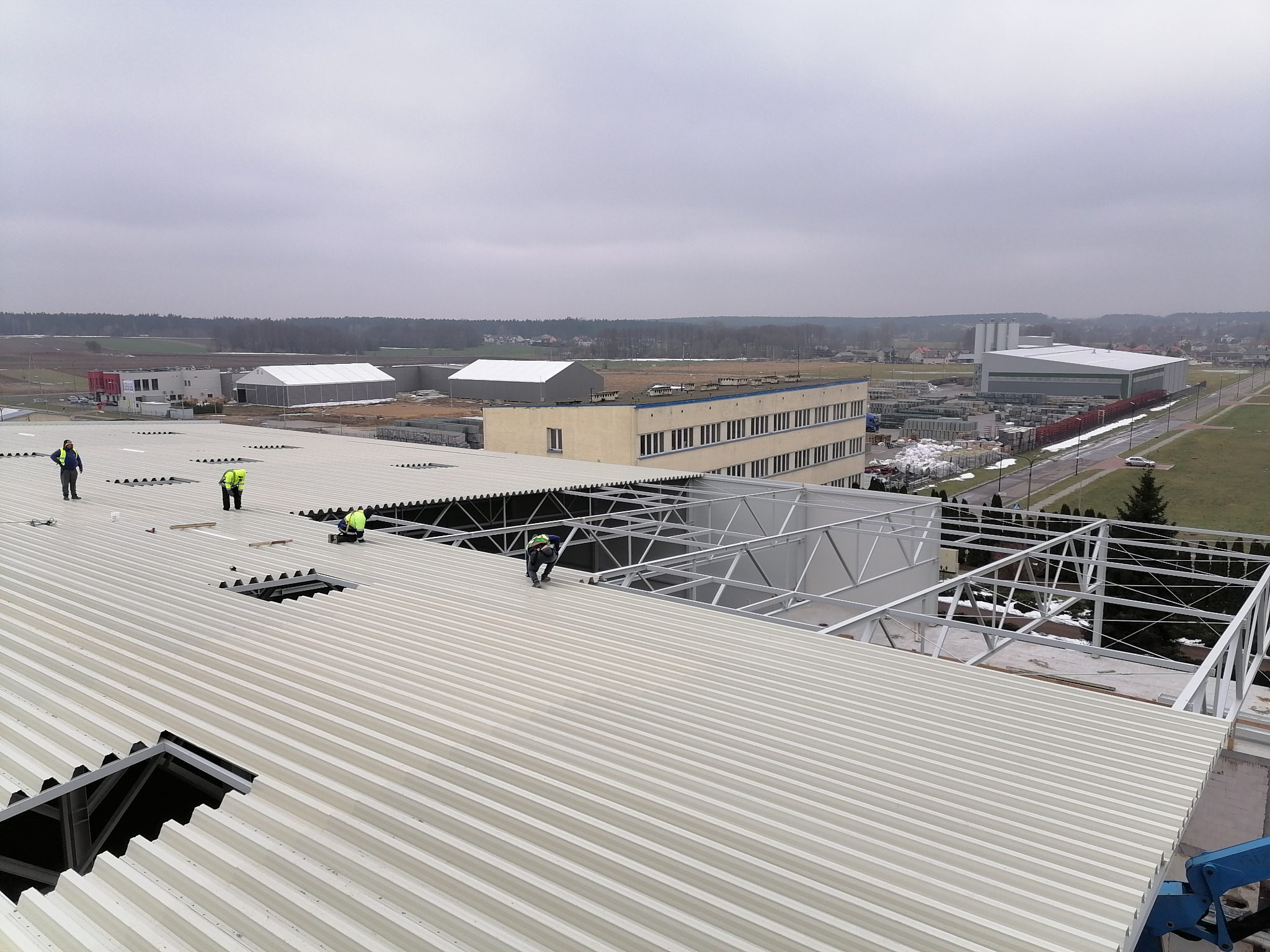 Pracownicy w kamizelkach odblaskowych montują konstrukcję na dachu z blachy trapezowej, widok z góry na budynki przemysłowe i tereny otwarte w tle.