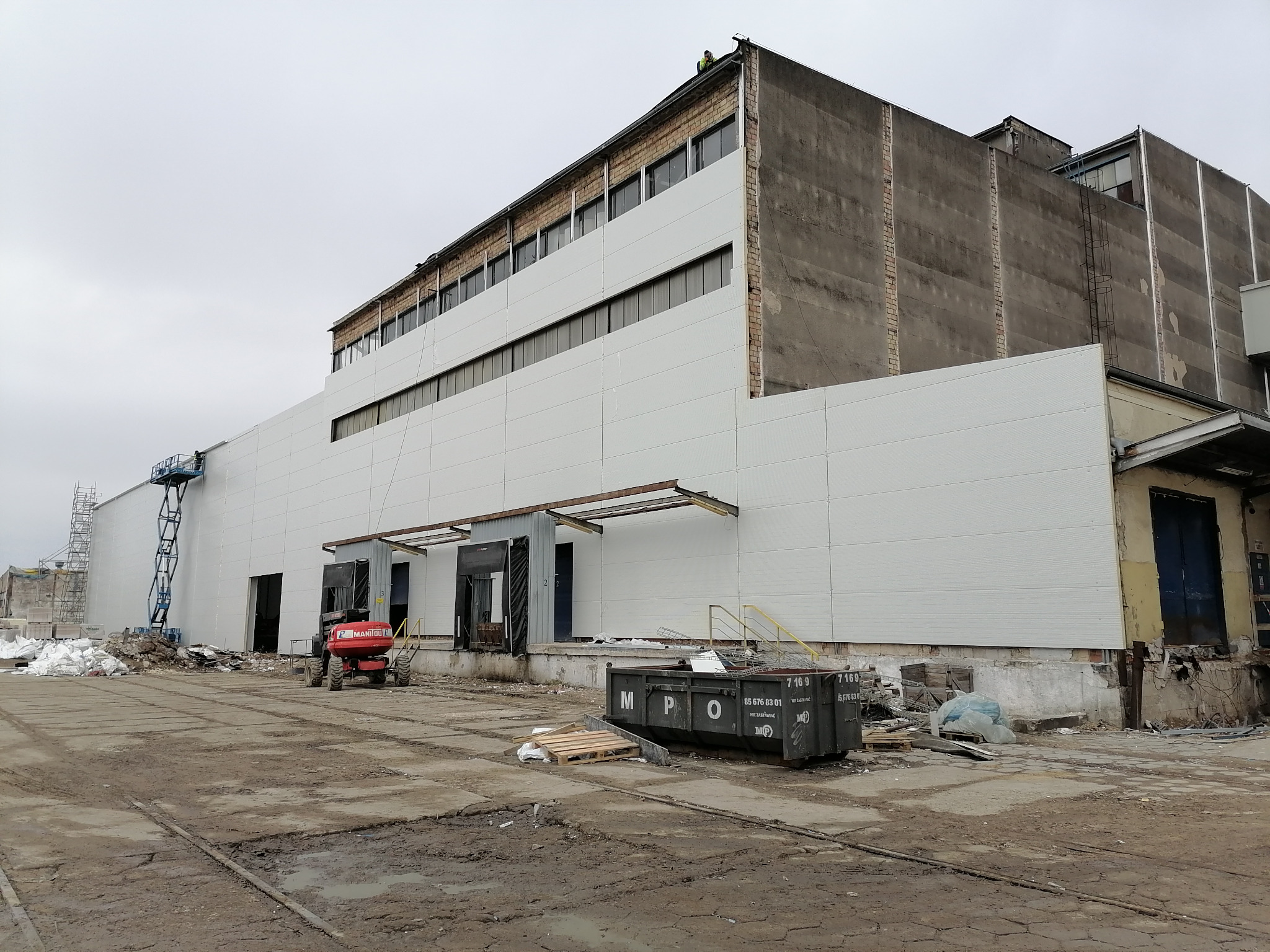 Montaż paneli na elewacji przemysłowego budynku, widoczny podnośnik koszowy i kontener MPO na błotnistym placu.