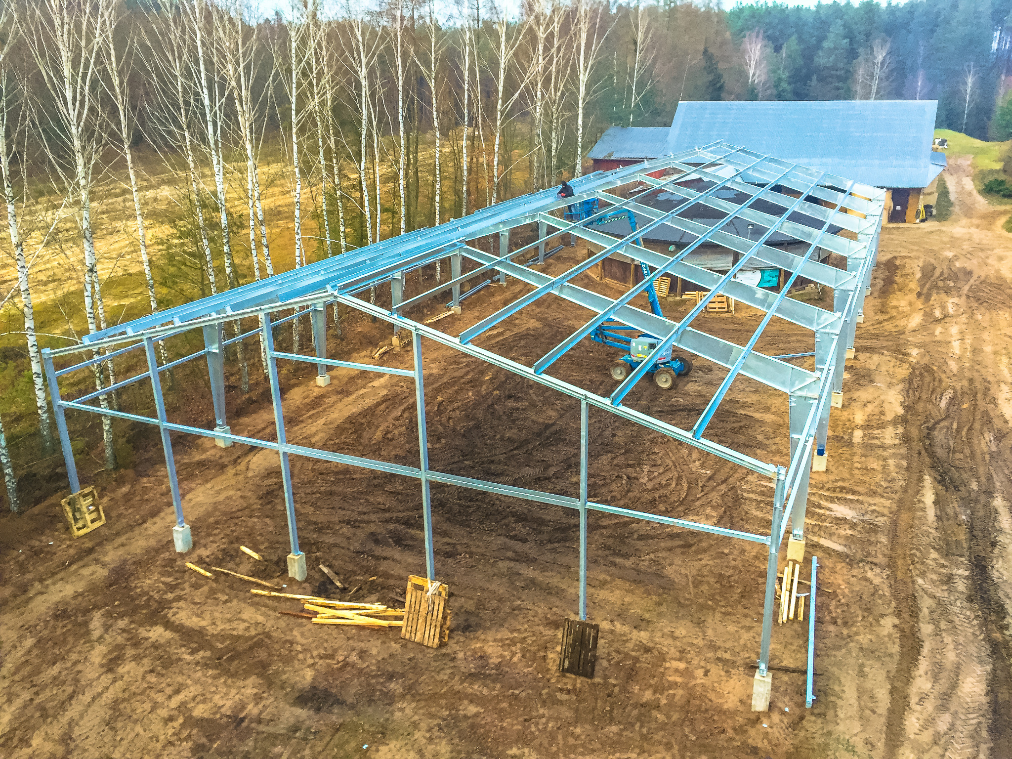 Wznoszenie metalowej konstrukcji budynku gospodarczego na fundamencie, widok z góry, w tle budynek gospodarczy i brzozowy zagajnik.