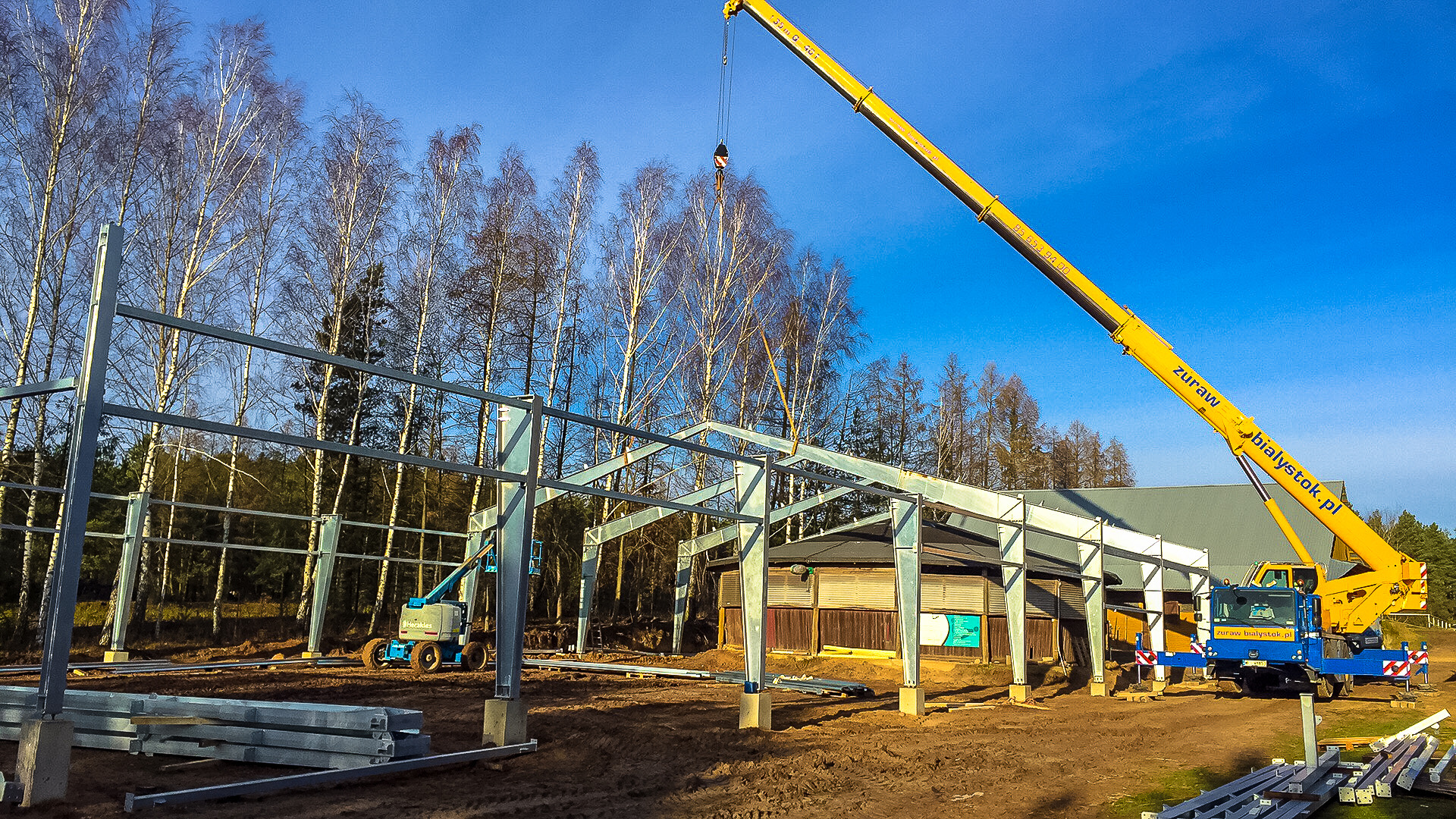 Montaż stalowej konstrukcji hali z użyciem dźwigu teleskopowego na tle brzozowego lasu i błękitnego nieba.