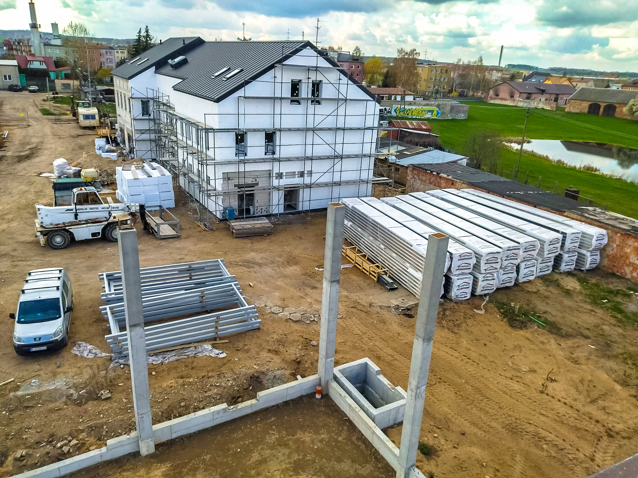 Budowa dwukondygnacyjnego domu z białymi ścianami, czarnym dachem i rusztowaniami, widoczne elementy konstrukcyjne, materiały budowlane i pojazdy na placu budowy w słoneczny dzień.