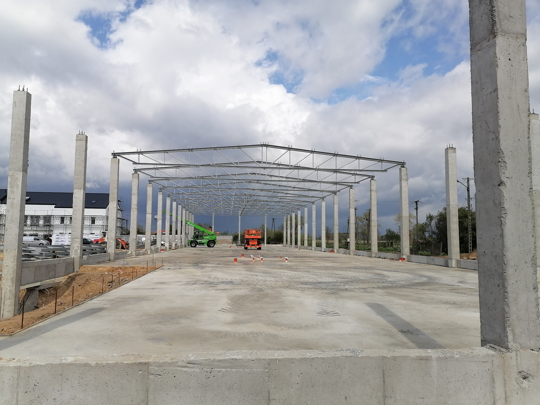 W trakcie budowy hali z prefabrykatów w Białymstoku: widoczne betonowe słupy, stalowa konstrukcja dachu, oraz maszyny budowlane na placu budowy.