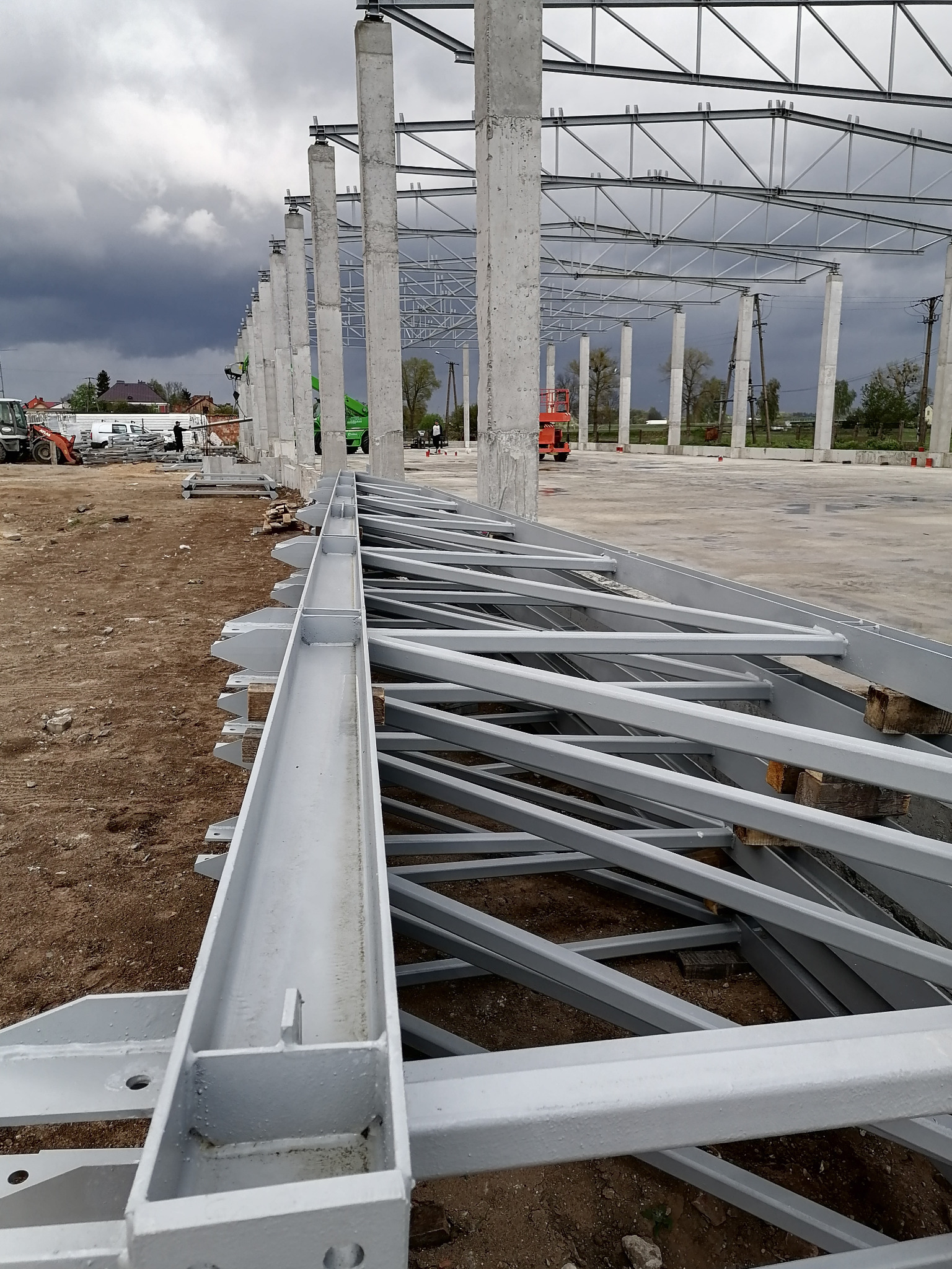 Konstrukcja stalowa hali w trakcie montażu, widoczne słupy betonowe, elementy kratownic i sprzęt budowlany w tle, pochmurne niebo.