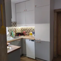 Szare i białe szafki kuchenne z miedzianymi uchwytami, wzorzyste płytki nad blatem, widoczny fragment drewnianej podłogi i drzwi