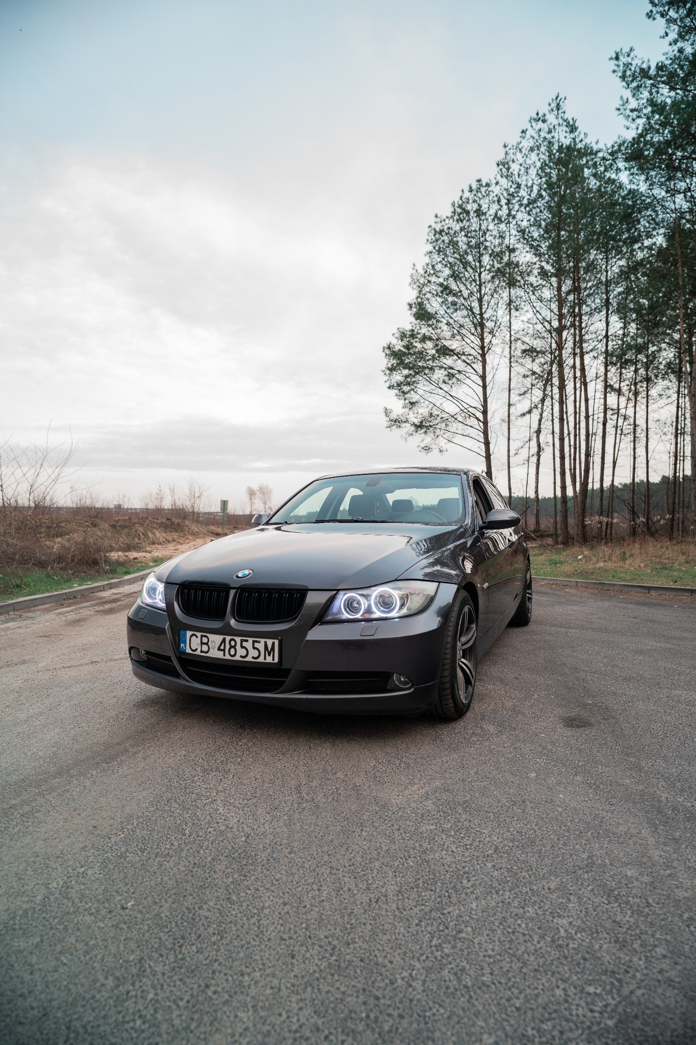 Szary samochód BMW serii 3 E90 z charakterystycznymi ringami LED w reflektorach, zaparkowany na asfaltowej drodze w pobliżu lasu.
