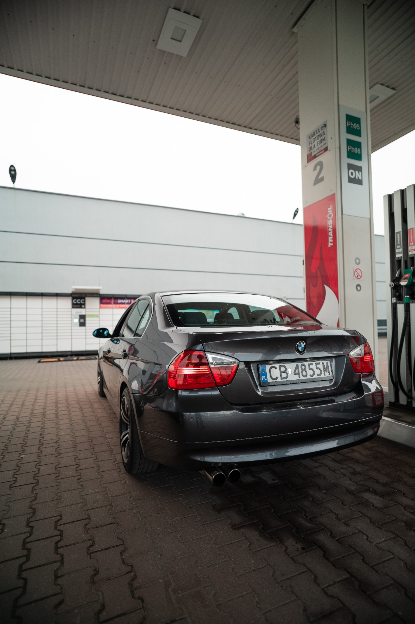 Szary samochód BMW serii 3 (E90) tankujący na stacji benzynowej, widok z tyłu, tablica rejestracyjna CB 4855M, pod dystrybutorem paliwa z oznaczeniami Pb95, Pb98 i ON.