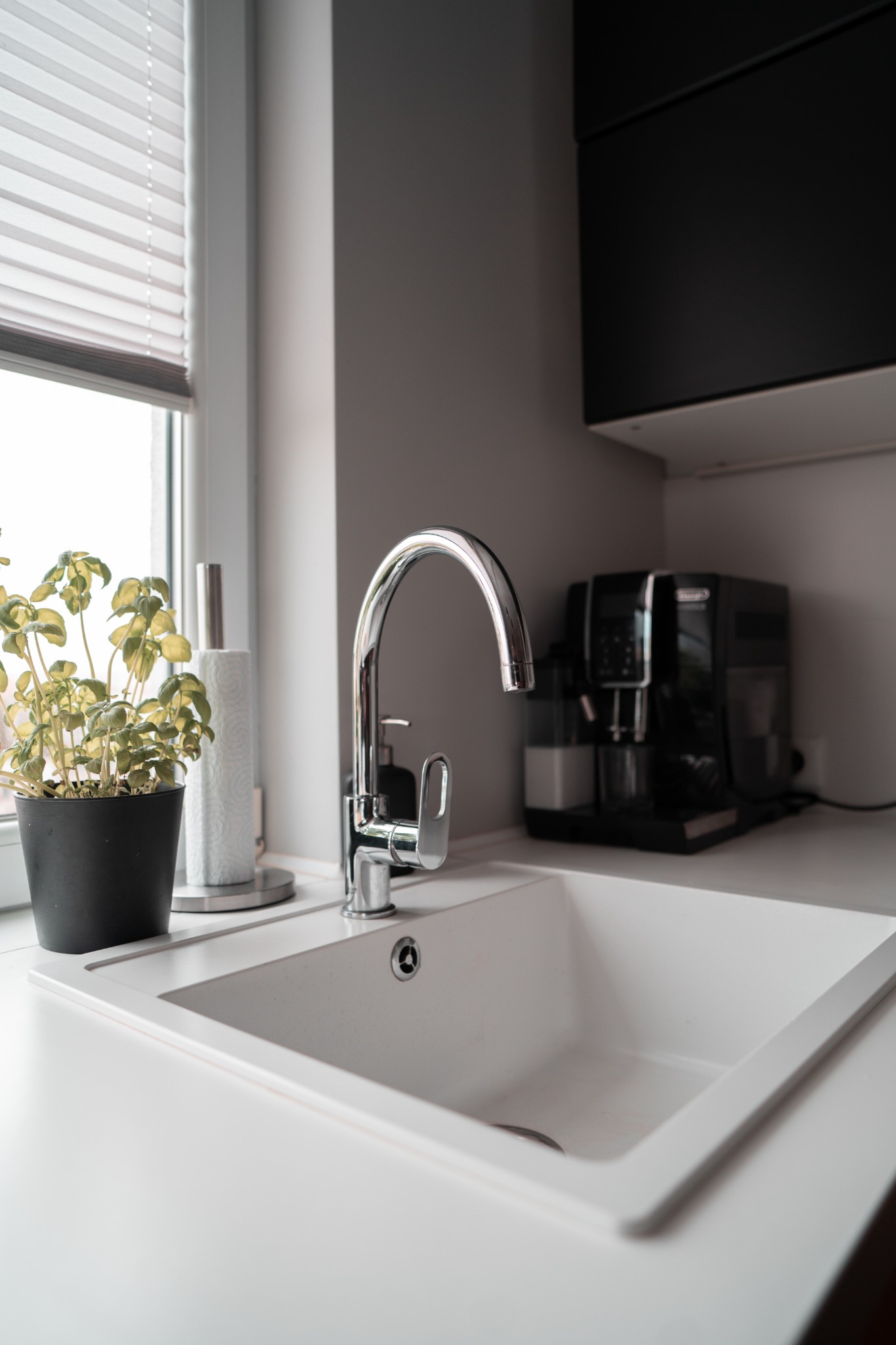 Biały zlewozmywak z chromowaną baterią, obok doniczka z ziołami i ekspres do kawy w tle, oświetlone przez okno z żaluzjami.