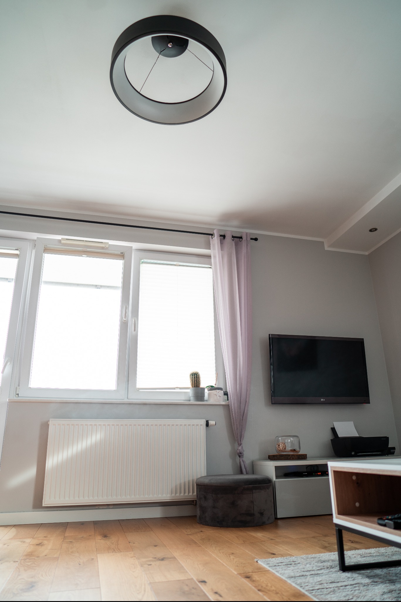 Jasne wnętrze pokoju z drewnianą podłogą, białym grzejnikiem, oknem z roletami i fioletową zasłoną, telewizorem na ścianie, okrągłą lampą sufitową i szarym pufem