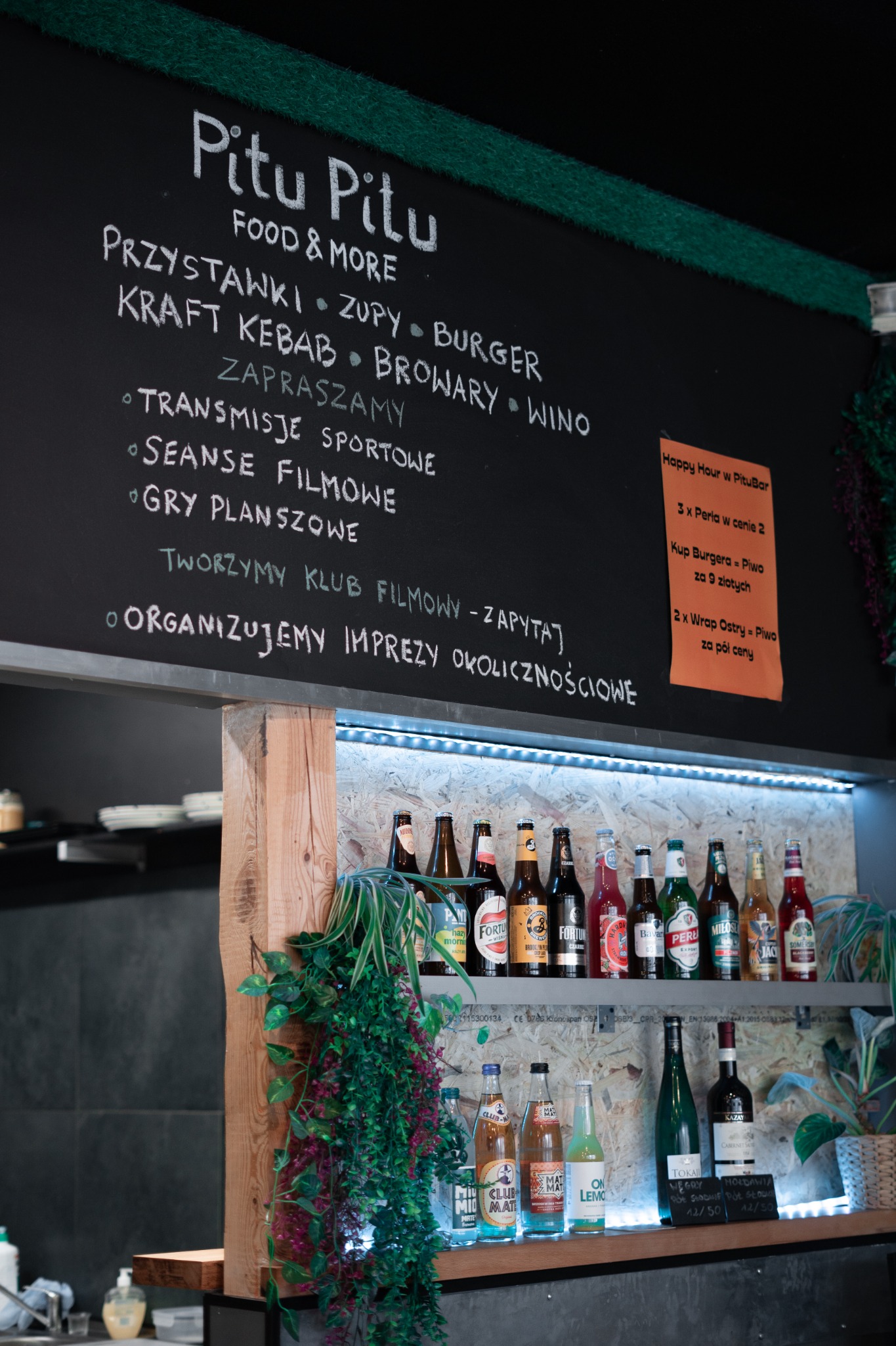 Wnętrze baru Pitu Pitu z menu wypisanym kredą na tablicy, butelki piwa i napojów na półkach podświetlonych LED, dekoracje roślinne.