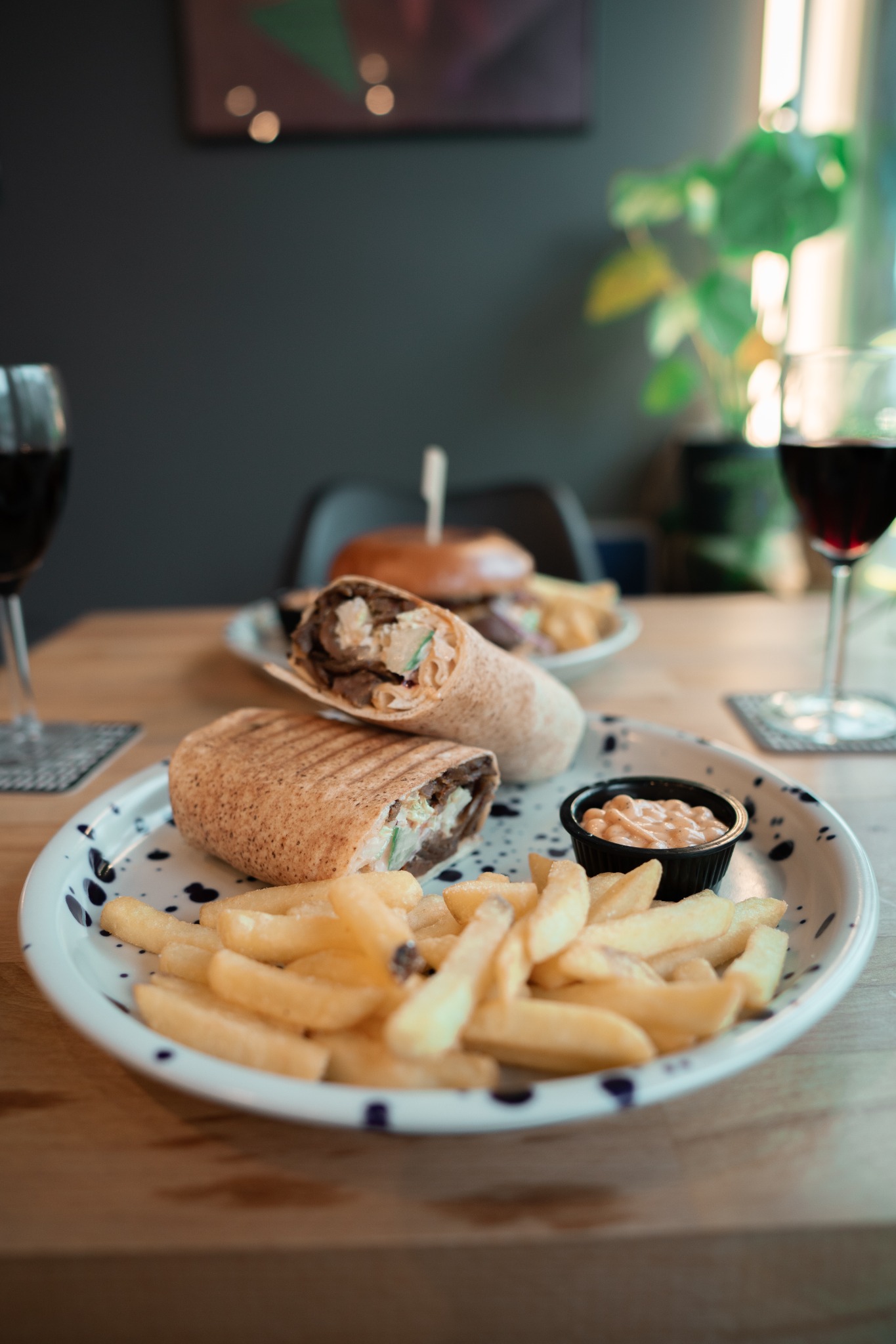 Dwa kawałki tortilli z mięsem i warzywami, frytki oraz sos na talerzu z niebieskimi kropkami, w tle burger i kieliszki z czerwonym winem.