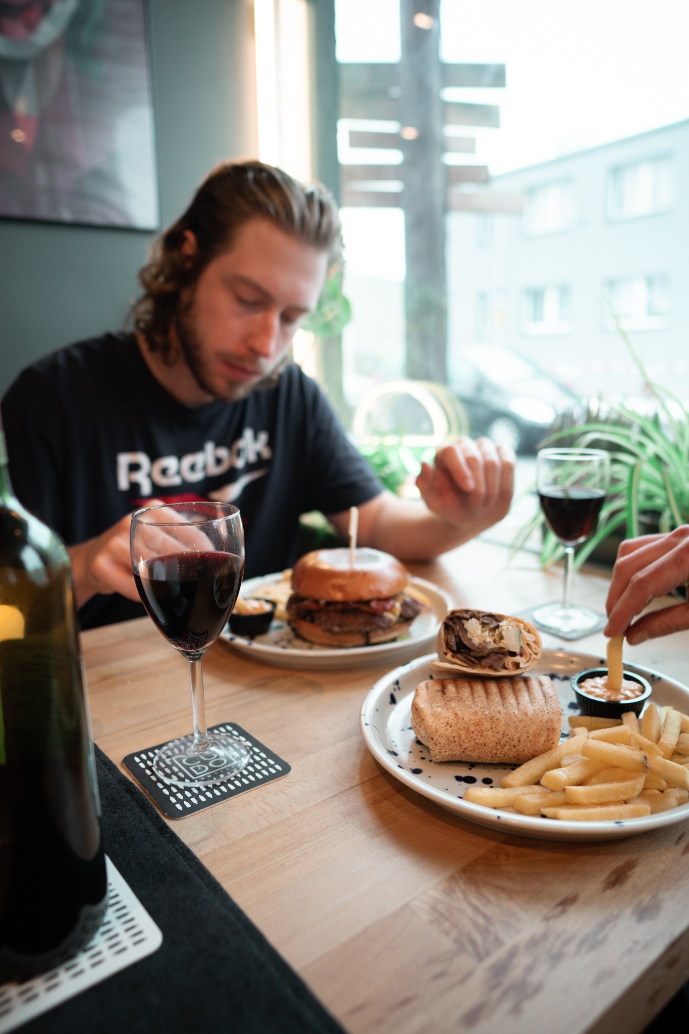Mężczyzna w czarnej koszulce z logo Reebok siedzi przy stole z burgerem, tortillą z mięsem, frytkami i czerwonym winem, w tle okno i roślina doniczkowa.